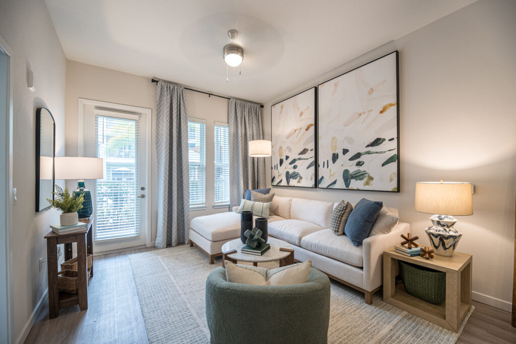 Modern living room with a white sectional sofa, large abstract art on the wall, a green accent chair, soft lighting from lamps, a small console table, and large windows with sheer curtains letting in natural light.