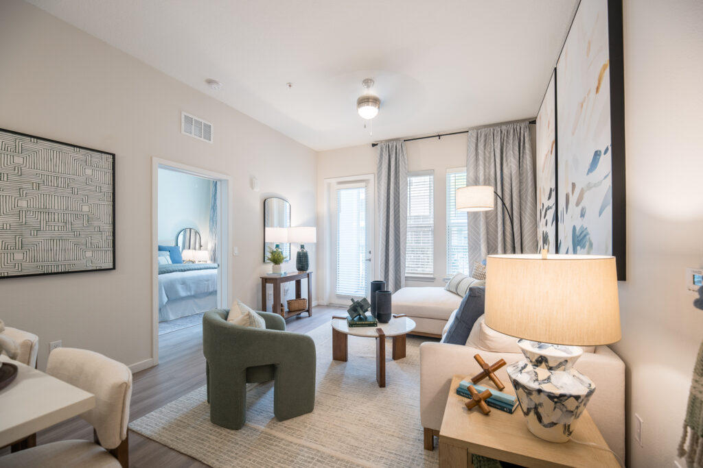 Modern living room with neutral tones, featuring a beige sofa, green armchair, coffee table, floor lamp, abstract art, and large windows with gray curtains; a bedroom is visible through an open doorway.