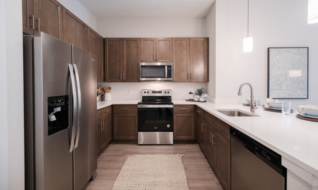 Modern kitchen with stainless steel appliances, brown wooden cabinets, white countertops, and a sink with a gooseneck faucet. A beige rug is centered on the floor, and pendant lights hang from the ceiling.