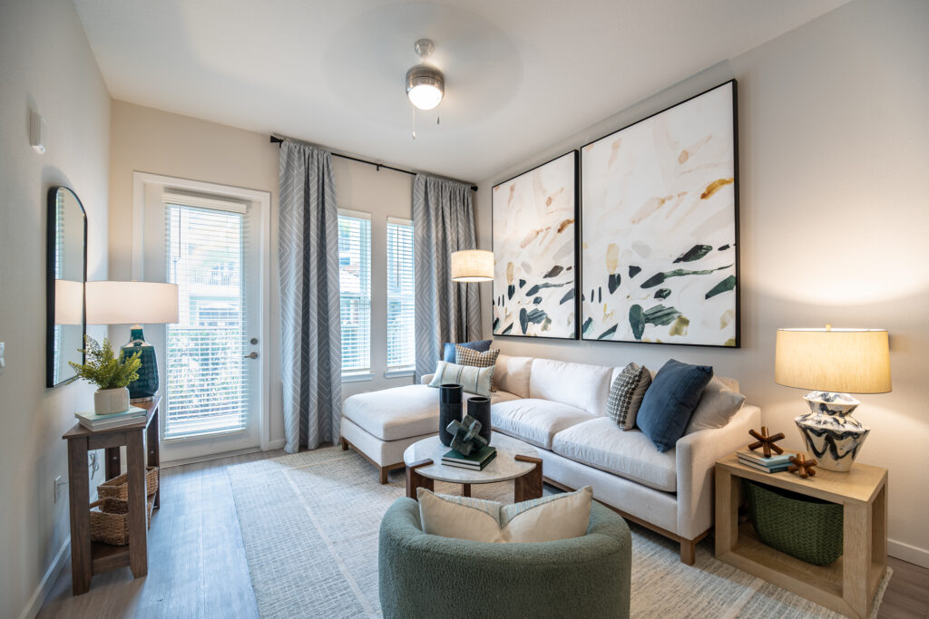 A modern living room with a light beige sectional sofa, abstract art on the wall, a floor lamp, side tables with lamps, and large windows with sheer curtains letting in natural light.