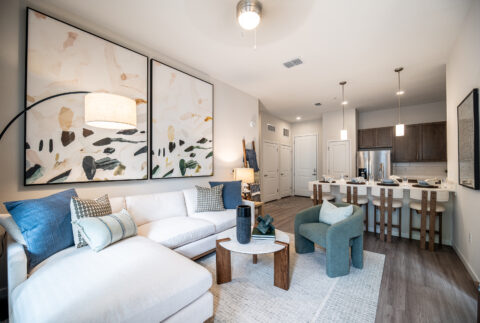 Modern living room with a white sectional sofa, blue and green accent pillows, a green armchair, abstract wall art, a small coffee table, and an open kitchen with wooden cabinets and bar stools in the background.