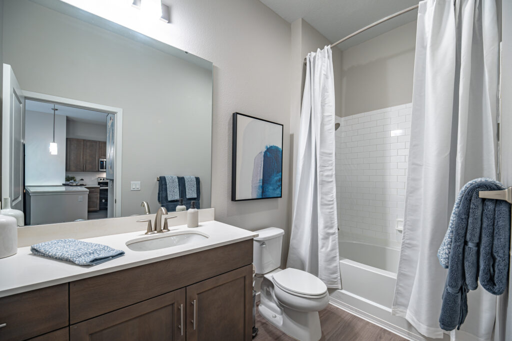 Modern bathroom with a wood vanity, white countertop, and sink. A toilet and bathtub with a white shower curtain are visible. Blue towels, abstract wall art, and a mirror add decorative touches. Doorway leads to a kitchen area.