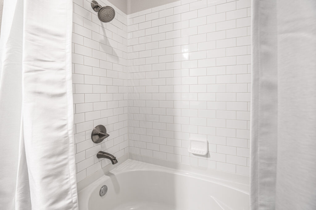 A clean white bathtub with a white subway tile wall, silver showerhead, faucet, drain, and a soap holder. White shower curtains are partially closed on both sides.