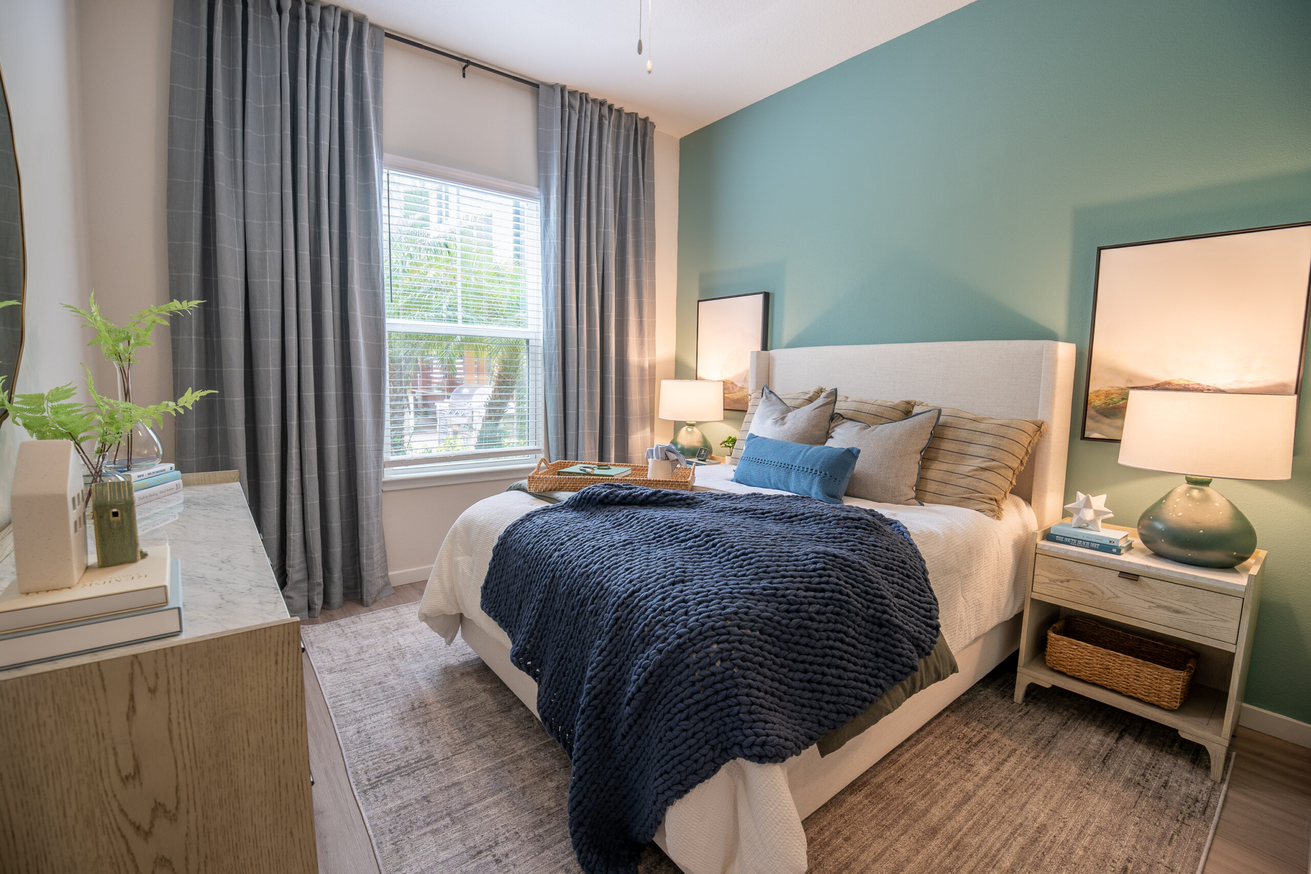 A cozy bedroom with a bed covered in white and navy blue bedding, two nightstands with lamps, a large window with gray curtains, green accent wall, and a dresser with decor and books. Natural light brightens the room.