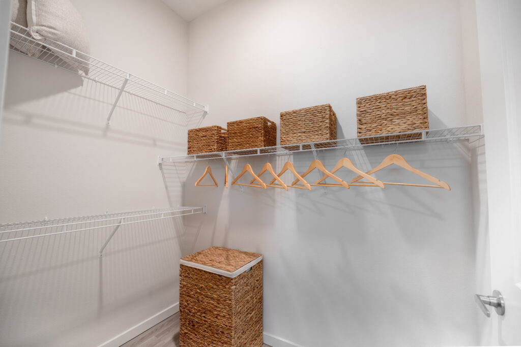 A walk-in closet with white walls, metal wire shelves, wooden hangers, woven storage baskets, and a matching woven laundry hamper on the floor. The closet is organized and minimalistic.