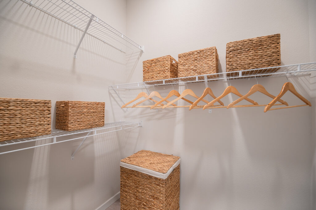 A clean, empty walk-in closet with white walls, wire shelves, several wicker storage baskets, wooden hangers, and a tall wicker hamper on the floor.