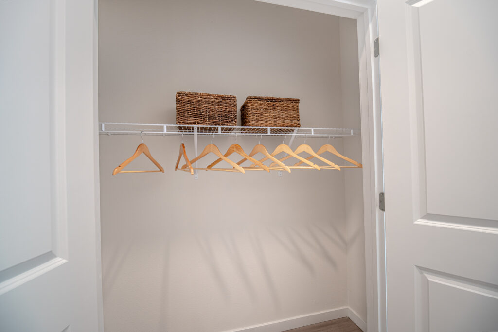 A small, empty closet with white walls, an open white door, a wire shelf holding two woven baskets, and several wooden hangers hanging from the shelf. The floor is light-colored.