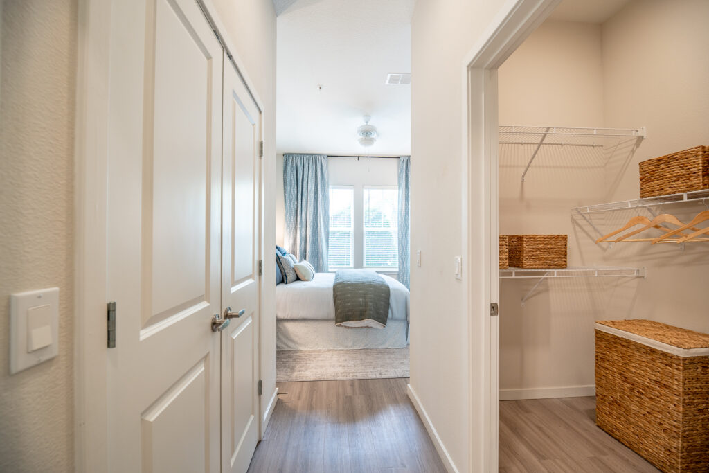View from a hallway into a bright bedroom with a neatly made bed, large windows with curtains, and an adjacent walk-in closet featuring wire shelves, hangers, and woven storage baskets.