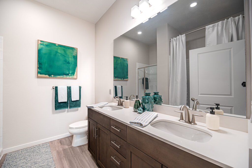 Modern bathroom with double sinks, brown cabinets, a large mirror, white countertop, and brushed metal faucets. A toilet and green artwork are visible, with folded towels hanging nearby and decorative items on the counter.