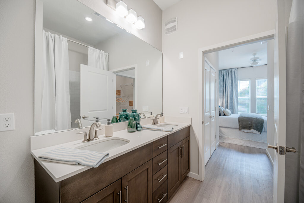 Modern bathroom with a double sink vanity, large mirror, decorative green glass bottles, and striped towels; view extends into a bright bedroom with a made bed and large windows.