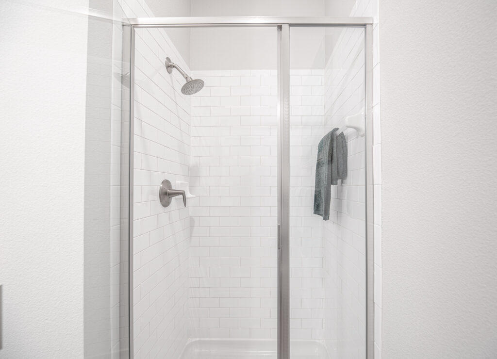 A modern glass shower with white subway tile walls, a silver showerhead, and a metal towel rack holding a folded gray towel. The space appears clean and minimalist.
