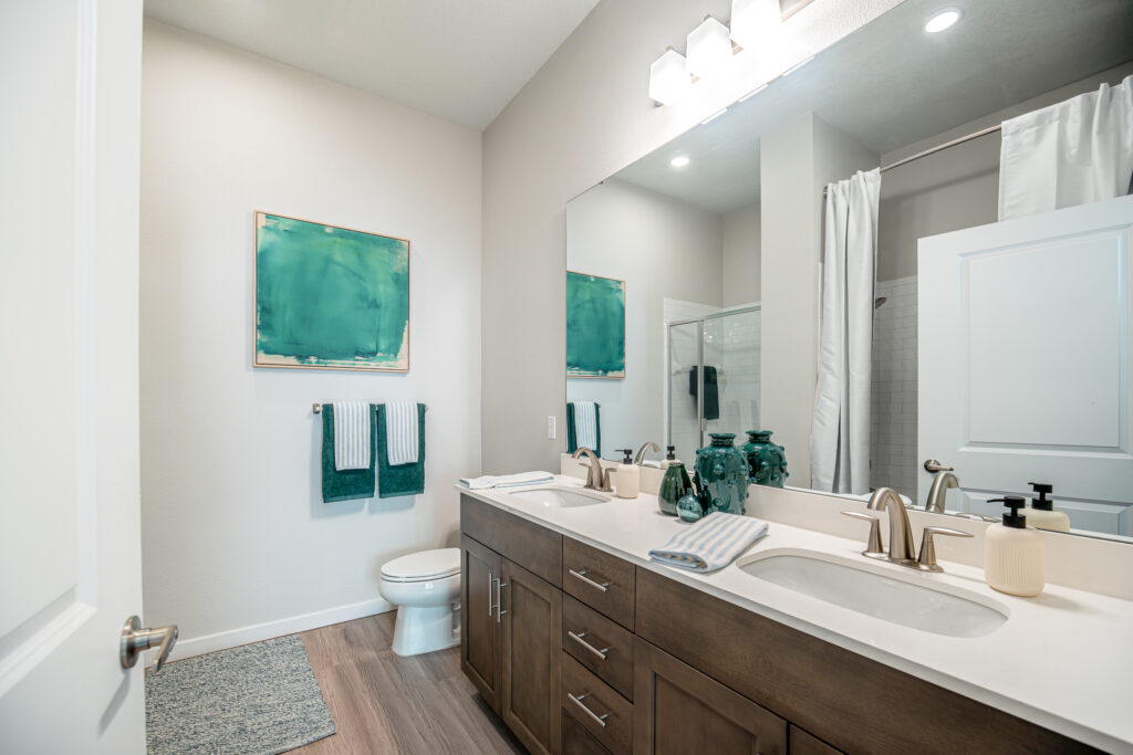Modern bathroom with double sinks, wood vanity, large mirror, and brushed nickel fixtures. Green artwork and towels add color. Toilet and shower with white curtain are visible; space is clean and bright.