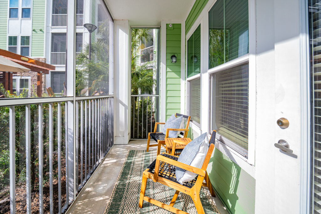 A sunny apartment balcony with two wooden chairs and cushions, a green wall, outdoor rug, and metal railing. Plants and neighboring buildings are visible outside the screened-in porch area.