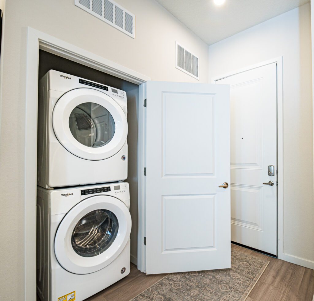 A white stacked washer and dryer set is tucked into a small laundry closet with an open double door next to a white entry door in a bright hallway with a patterned rug.