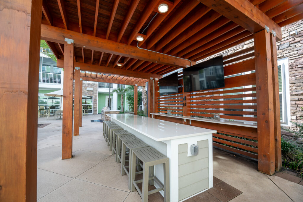 Outdoor covered patio area with a long white bar table, several stools, two large TVs mounted on a wooden slatted wall, and modern architecture visible in the background.