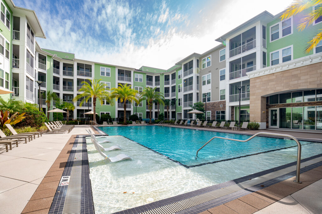 Modern apartment complex courtyard with a large outdoor swimming pool, lounge chairs, umbrellas, palm trees, and surrounding multi-story green and beige buildings under a partly cloudy sky.