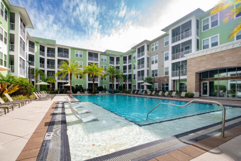 Modern apartment complex courtyard with a large outdoor swimming pool, lounge chairs, umbrellas, palm trees, and surrounding multi-story green and beige buildings under a partly cloudy sky.