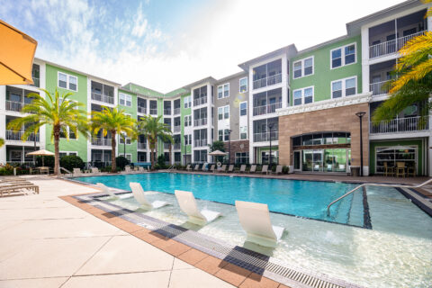 Modern apartment complex with green and white exterior, featuring a large outdoor swimming pool, lounge chairs, palm trees, and a spacious pool deck under a bright, sunny sky.