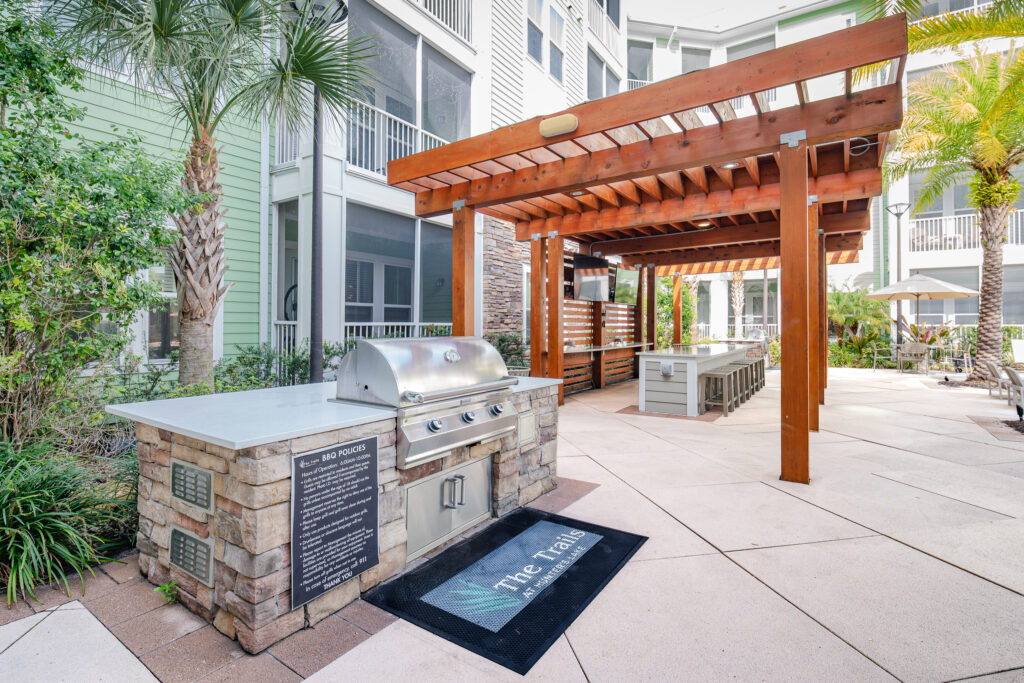 Outdoor grilling area with a stainless steel barbecue grill built into a stone island, under a wooden pergola, surrounded by tropical plants and patio seating in an apartment courtyard. The Trail is printed on a doormat.