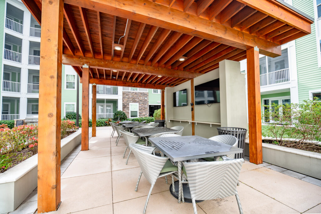 Outdoor patio with a wooden pergola, several metal tables and chairs, a wall-mounted TV, and surrounding modern apartment buildings with balconies and landscaped greenery.