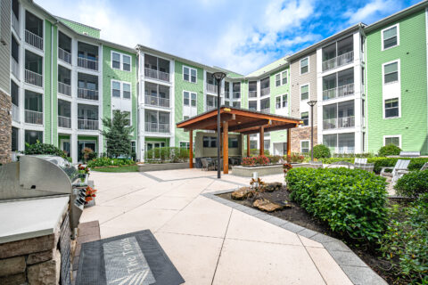 A modern apartment building with green siding surrounds a landscaped courtyard featuring a wooden pergola, outdoor seating, a grill area, plants, and a pathway under a partly cloudy sky.