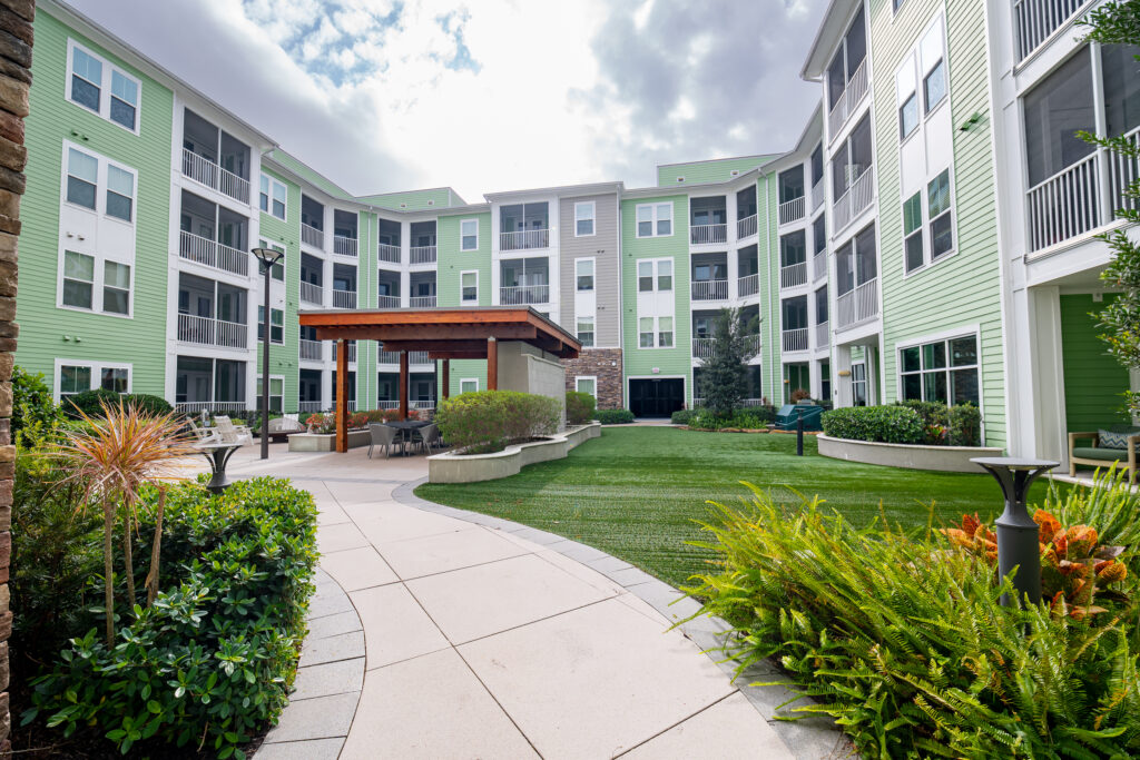 A modern apartment complex courtyard with green grass, lush landscaping, outdoor seating under a wooden pergola, and four-story green and white residential buildings surrounding the area.