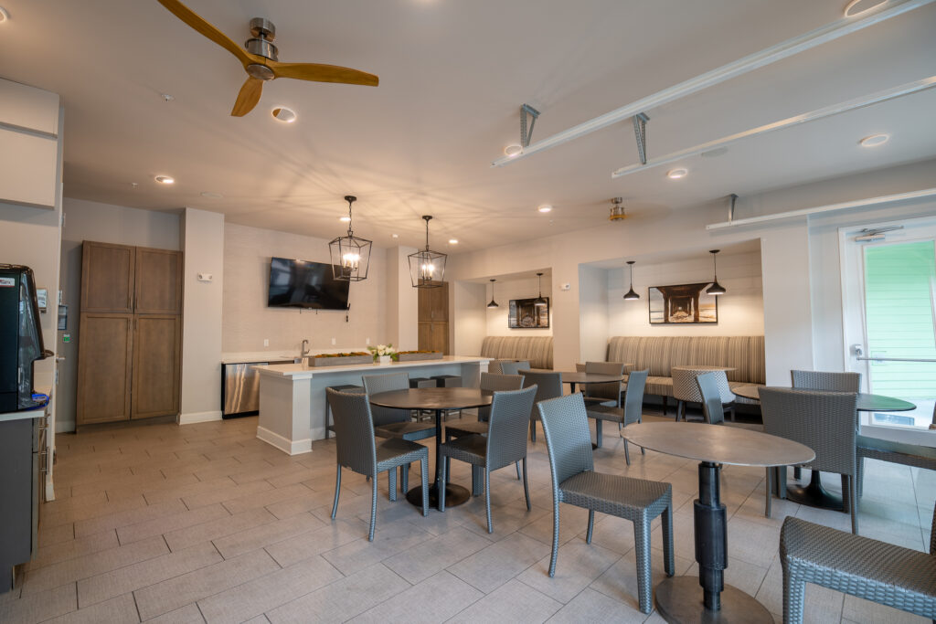 Modern communal lounge with gray tile flooring, round tables, woven chairs, a kitchenette with a TV above, pendant lights, and booth seating along the wall. Ceiling fans and natural light create an inviting atmosphere.