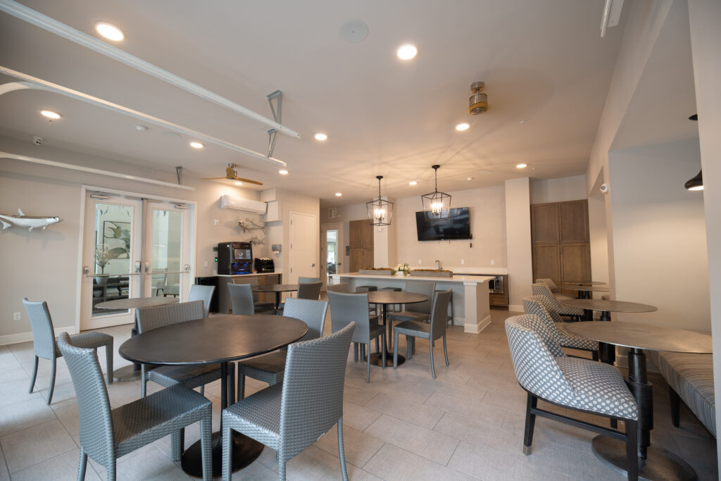 A modern, well-lit lounge area with round tables and chairs, a kitchenette with barstools, a coffee station, wall-mounted TV, and décor featuring shark sculptures on the light-colored walls.