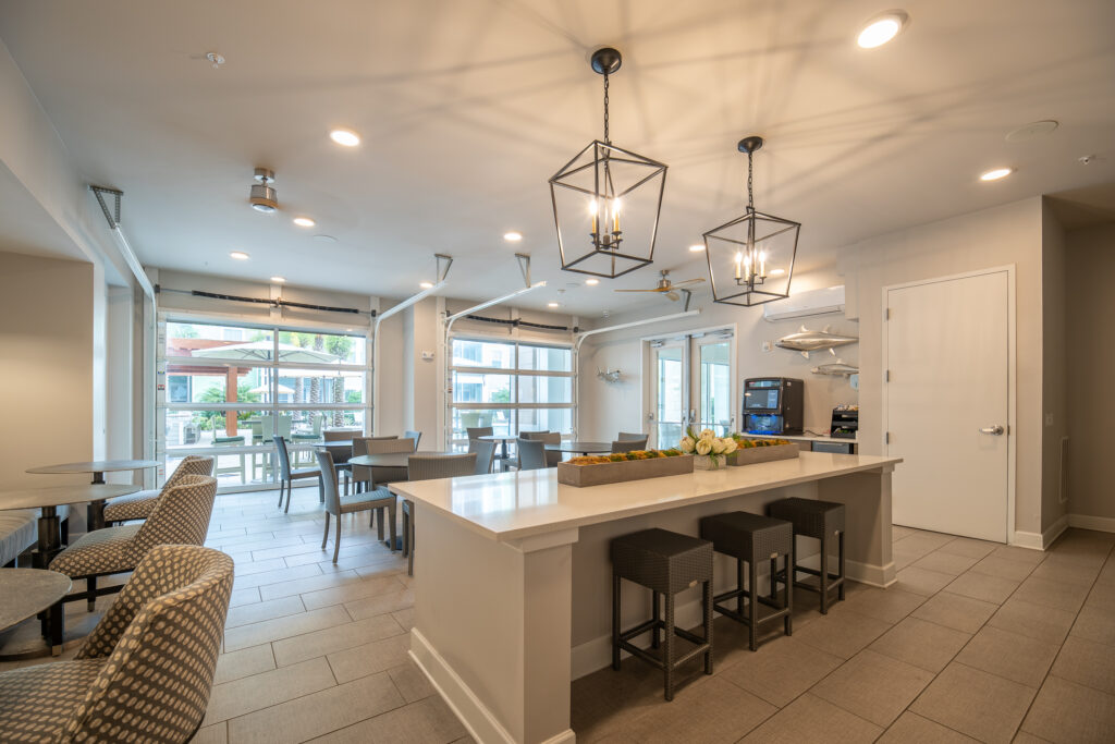 Modern communal kitchen and dining area with a large island, barstools, patterned chairs, dining tables, pendant lights, and large windows letting in natural light. Contemporary decor with neutral tones.