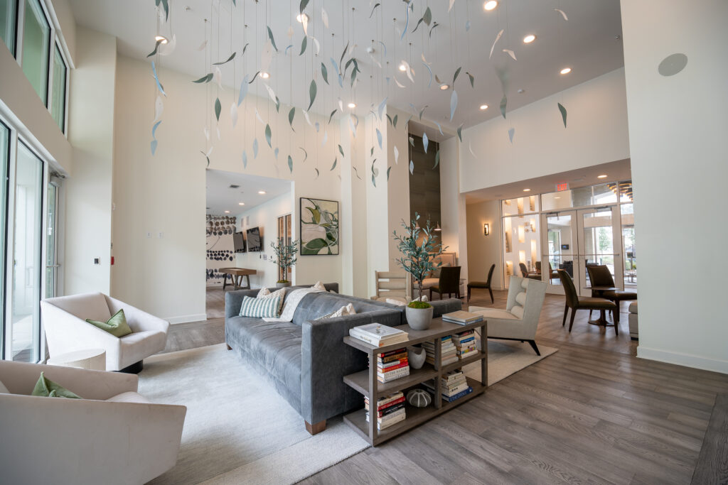 A modern, airy lounge with a gray sofa, armchairs, bookshelves, and hanging leaf decorations. Large windows bring in natural light; dining and seating areas are visible in the background.