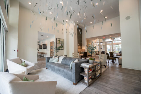 Modern, spacious lounge with a gray sofa, two white armchairs, and side tables with books and plants. Leaf-like decorations hang from the ceiling; large windows let in natural light, creating an inviting, open atmosphere.