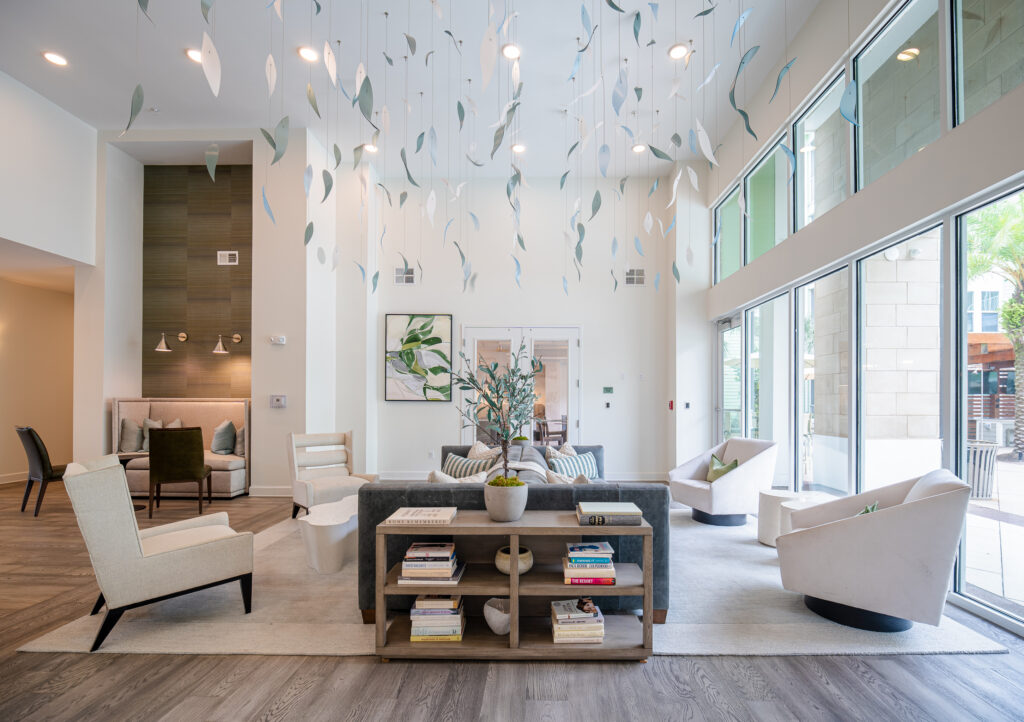 Modern, bright lounge with large windows, stylish armchairs, a sofa, bookshelves, and decorative hanging art pieces from the ceiling. Natural light fills the space, highlighting contemporary decor and green plants.