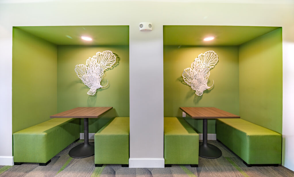 Two green booth seating areas with wooden tables and benches, separated by a white wall. Each booth has a decorative white wall sculpture shaped like abstract wings and is lit by a ceiling light.