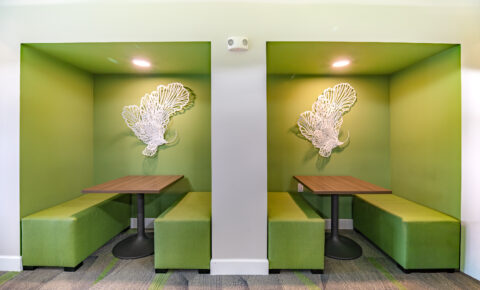 Two green booth seating areas with wooden tables and benches, separated by a white wall. Each booth has a decorative white wall sculpture shaped like abstract wings and is lit by a ceiling light.