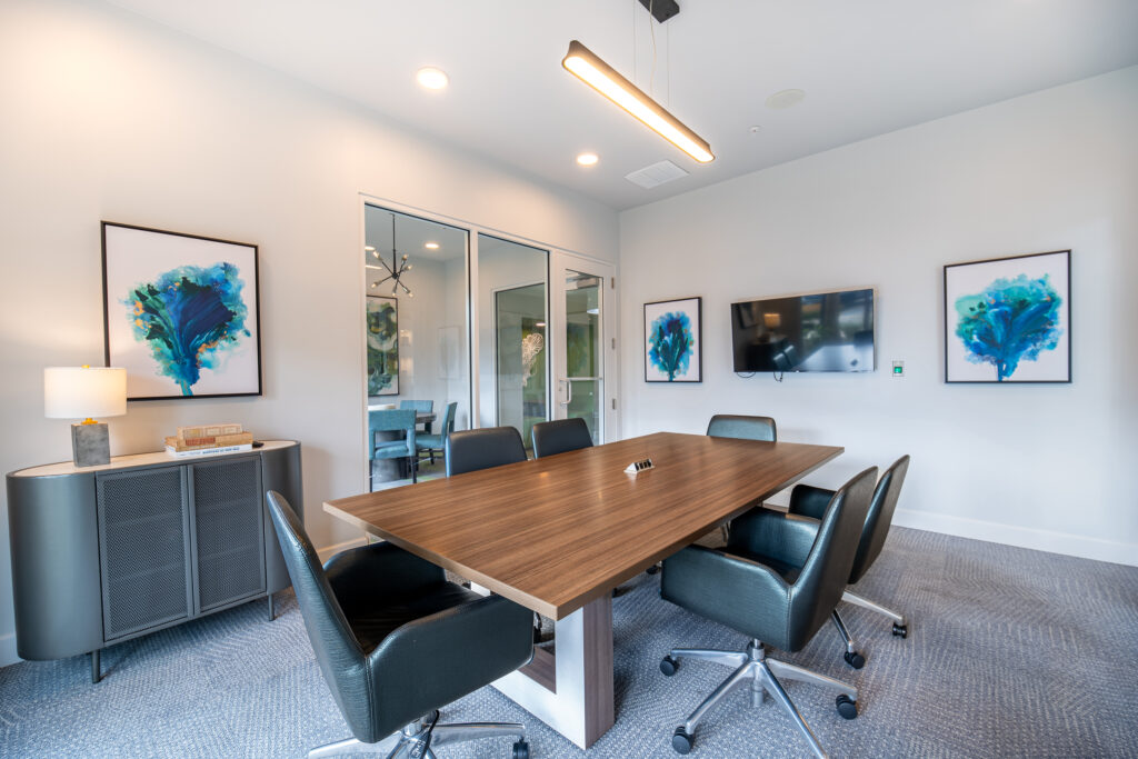 Modern conference room with a rectangular wooden table, six black chairs, wall-mounted TV, abstract art on the walls, glass doors, and a sideboard with a lamp and books. The space is well-lit and neatly organized.