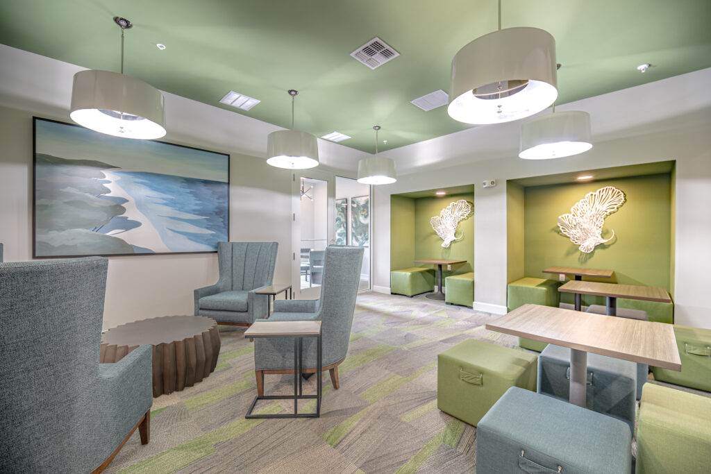 A modern lounge with green and gray decor, soft seating, small tables, pendant lights, abstract beach painting, and shell wall art, featuring a relaxed and inviting atmosphere.